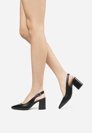 Zapatos de tacón negro con punta afilada, tacón grueso y correa ajustable en el tobillo; textura de cuero suave con un diseño minimalista.