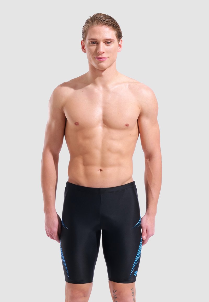 Young muscular man standing wearing black swim jammers with blue dotted patterns on sides against a plain background.