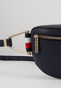 Borsa a tracolla in pelle blu navy con hardware dorato, chiusura con zip e un accento in tessuto a righe rosso, bianco e blu. Forma rotonda.