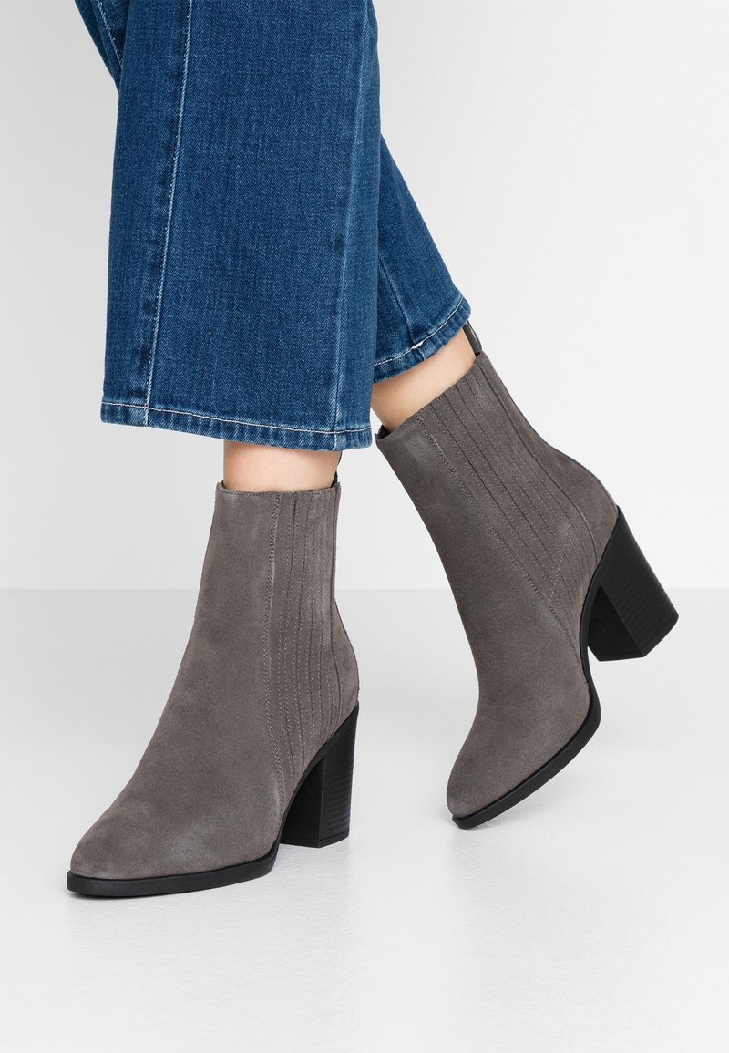 Bottes grises en daim montant à la cheville avec talons carrés portées avec un jean bleu raccourci sur fond blanc.
