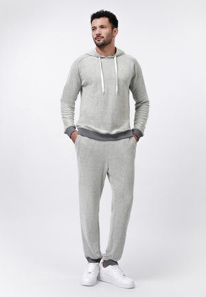 Lichtgrijze gebreide hoodie met witte trekkoorden, gecombineerd met bijpassende joggers met grijze boorden. Draagt witte sneakers.