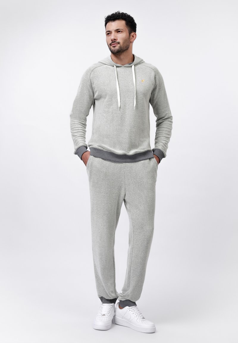 Helle graue, strukturierte Kapuzenjacke mit weißen Kordeln, kombiniert mit passenden Jogginghosen mit grauen Bündchen. Getragen mit weißen Sneakers.