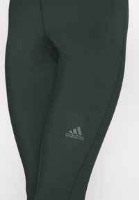 Tmavě zelené legíny vyrobené z pružného materiálu s hladkou texturou, s decentním šedým logem Adidas blízko spodní části nohy.