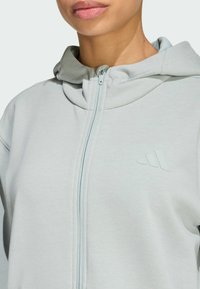 Vaaleanharmaa vetoketjuhuppari, jossa on teksturoitu kangas, korkea kaulus ja hillitty logo rinnassa, korostaen minimalistista muotoilua.