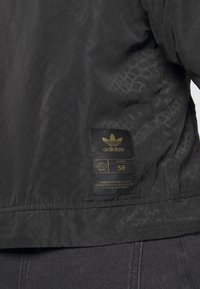 Svart jacka med ett subtilt texturerat mönster, med en rektangulär etikett med guld detaljer och Adidas-logotyp, storlek 50.