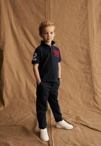 Tamsiai mėlynas polo marškinėliai su raudonu siuvinėjimu "SUPERDRY STATE ATHLETE" tekstu, numeris "2" ant rankovės, derinami su tamsiomis kelnėmis ir baltais sportiniais bateliais.