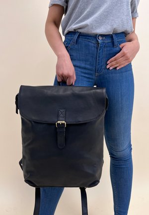 Svart läderryggsäck med klaffstängning, guldbucklesaccent och justerbara remmar, hålls av en person som bär blå jeans och en grå skjorta.