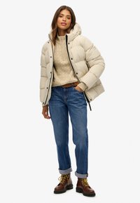 Beige dunjakke med hætte over en cremefarvet kabelstrikket sweater. Blå jeans med op-rullede manchetter og brune snørestøvler med gule detaljer.