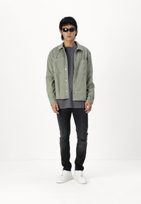Lys grønn knapp-up jakke over en mørkegrå skjorte, parret med svarte jeans og hvite joggesko. Modellen bruker svarte solbriller.