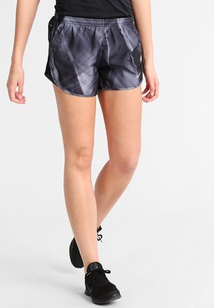 Osoba ubrana w czarne i szare sportowe szorty tie-dye, czarny top i czarne buty sportowe, idąca z jedną nogą wysuniętą do przodu na białym tle.