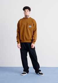 Sudadera marrón con cuello redondo, con un bolsillo en el pecho y detalle de logo. Combinada con pantalones negros y zapatillas negras.