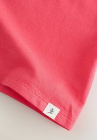 Camiseta coral con textura suave, que presenta una etiqueta blanca con un gráfico de hoja en el dobladillo. Diseño sencillo, mangas cortas y sin estampados.