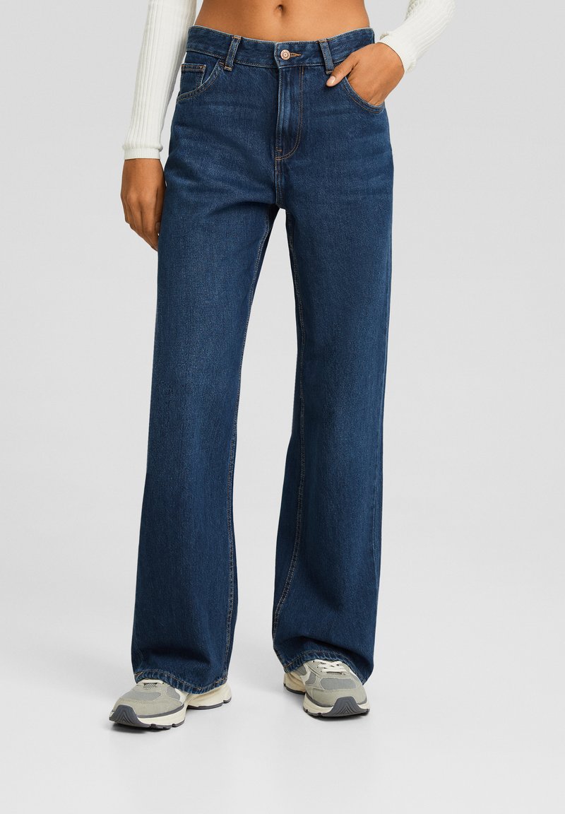 Jean en denim bleu foncé à jambes larges et taille haute, tissu texturé, avec des détails de coutures contrastants. Assorti à des baskets gris clair.