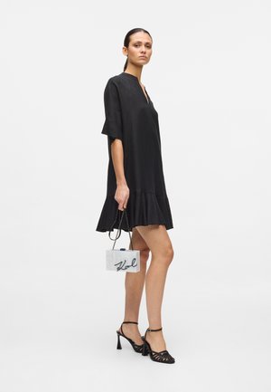 Vrouw in een loszittende zwarte jurk die een kleine witte, doosvormige tas met kettingriem vasthoudt, met zwarte sandalen met hoge hak en bandjes aan.