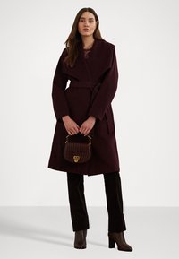 Cappotto in lana bordeaux con ampi revers e vita segnato da una cintura, abbinato a pantaloni in velluto marrone e una borsa testurizzata. Tessuto liscio, silhouette elegante.