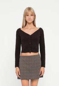 Svart cropped cardigan med V-hals, lange ermer og fem knapper foran; brukt sammen med et rutete miniskjørt i beige og brune toner.