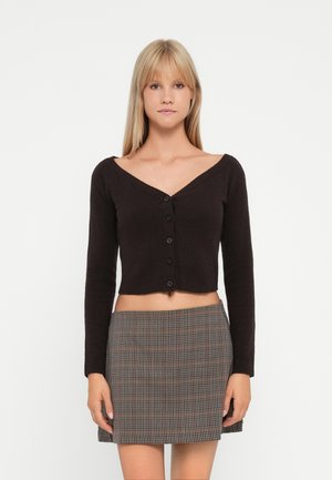 Zwarte cropped cardigan met een V-hals, lange mouwen en vijf voorste knopen; gedragen met een geruite minirok in beige en bruine tinten.