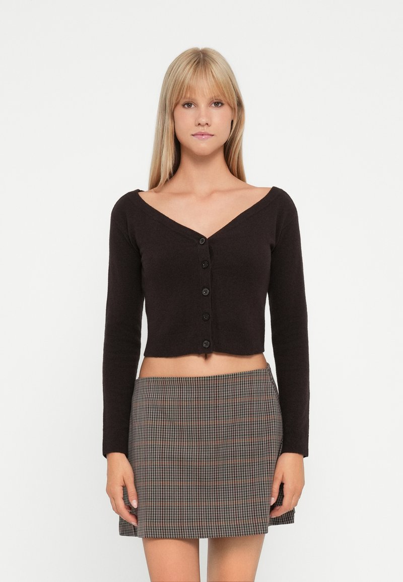 Svart cropped cardigan med V-hals, lange ermer og fem knapper foran; brukt sammen med et rutete miniskjørt i beige og brune toner.