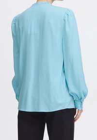 Blusa azzurro chiaro con maniche lunghe in tessuto testurizzato, maniche arricciate e un collo leggermente arruffato. Vestibilità casual con una caduta fluida.