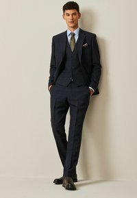 Costume à carreaux bleu marine avec une veste ajustée, un gilet et un pantalon. Accompagné d'une chemise bleu clair et d'une cravate à motifs. Chaussures noires.