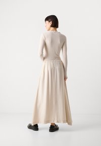 Long, beige dress with a ribbed textured top and a flowing, gathered skirt. The garment features a ruffled neckline and is paired with black shoes.