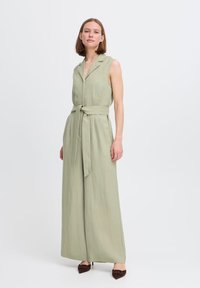 Salopette oliva senza maniche con colletto, vita con cintura, gambe larghe e finitura in tessuto testurizzato. Abbinata a scarpe marroni a punta.