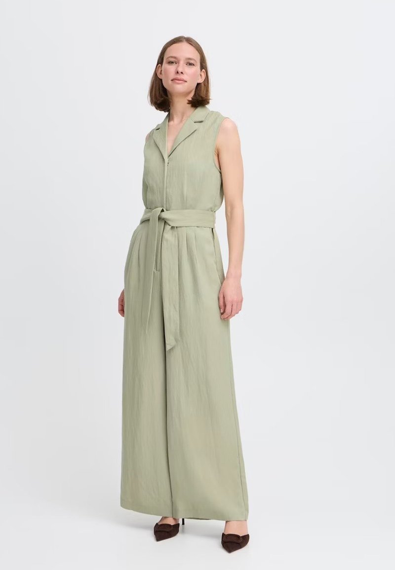 Salopette oliva senza maniche con colletto, vita con cintura, gambe larghe e finitura in tessuto testurizzato. Abbinata a scarpe marroni a punta.