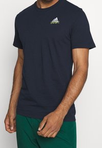 Námořnická modrá bavlněná tričko s krátkým rukávem, kulatým výstřihem a malým zeleným logem Adidas na hrudi. Měkká textura, standardní střih.