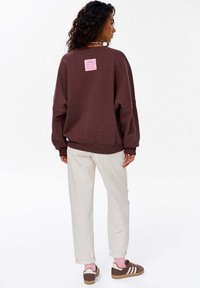 Hnědá oversized mikina s růžovou nášivkou na zádech, kombinovaná se světlými kalhotami a hnědými teniskami s bílými pruhy.