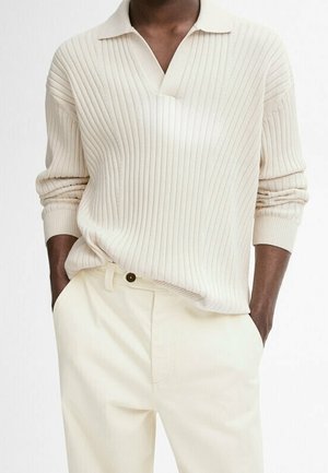 Mørkhudet person iført en cremefarvet ribstrikket sweater med krave og cremefarvede bukser med hænderne i lommerne.