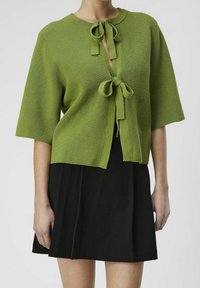 Femme portant un cardigan vert à manches courtes en maille avec deux nœuds à l'avant, sur une jupe noire, sur un fond uni.