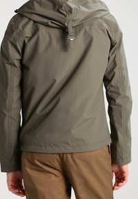 Uomo che indossa una giacca con cappuccio verde oliva e pantaloni marroni, visto da dietro, in piedi con le braccia rilassate lungo i fianchi.