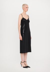 Schwarzes Midikleid mit dünnen Trägern, einem taillierten Oberteil und einem geraden, fließenden Saum. Der Stoff hat eine glänzende Textur. Getragen mit spitzen schwarzen Absätzen.