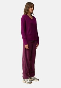 Sweter z dekoltem w serek, długim rękawem w głębokim fioletowym kolorze, w zestawieniu z szerokimi spodniami z kordurowej tkaniny w dopasowanym kolorze. Model nosi wzorzyste trampki.