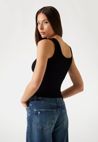 Černý žebrovaný top s širokými ramínky, spárovaný s světle modrými roztrhanými džíny. Modelka je z profilu.