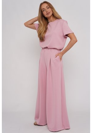 Kobieta z długimi blond włosami, ubrana w luźne, różowe spodnie z wysokim stanem i pasującą bluzkę z krótkim rękawem, stojąca z jedną ręką w kieszeni.