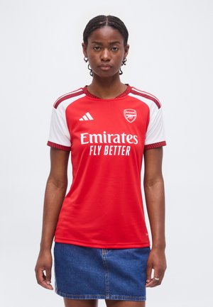 Joven mujer con cabello trenzado que lleva puesta la camiseta de fútbol roja y blanca del Arsenal y falda vaquera, de pie contra un fondo liso.