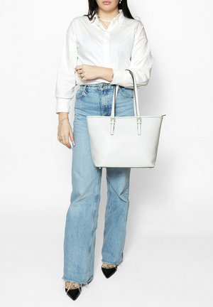 Sac fourre-tout blanc avec une surface texturée, deux poignées et des éléments en métal argenté. Le mannequin porte une chemise blanche et un jean bleu clair avec des jambes évasées.