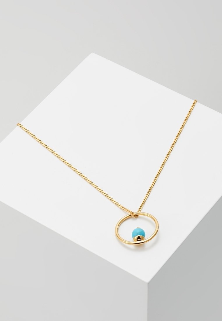 Collier en chaîne dorée avec un pendentif circulaire tenant une petite perle bleue, exposé sur une surface géométrique blanche.