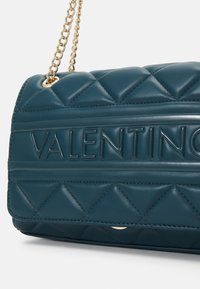 Zelena prešita usnjena torba s povišanim logotipom "VALENTINO", zlatim verižnim ročajem in teksturo v obliki diamanta na površini.