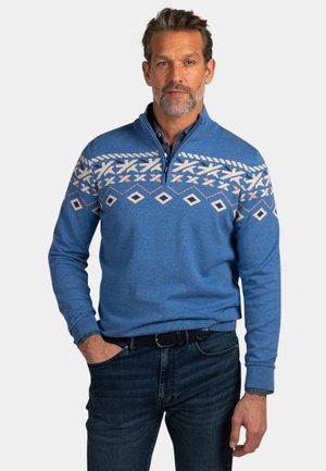 Man in een blauwe trui met een halve ritssluiting en witte geometrische patronen, staand met één hand in de spijkerbroekzak tegen een effen achtergrond.