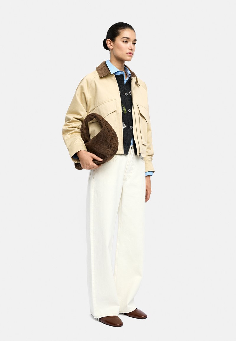 Kobieta ubrana w beżową kurtkę na czarnym swetrze i niebieskiej koszuli, w białe szerokie spodnie, brązowe buty, trzymająca torebkę w brązową strukturę.