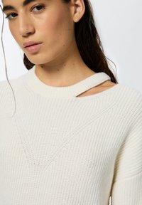 Maglione bianco in lavorazione a maglia con texture a coste, scollatura a girocollo con dettaglio a taglio sulla spalla e motivi geometrici di cucitura.