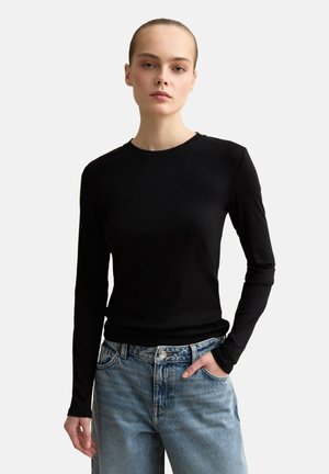Frau mit zurückgekämmtem Haar, die ein eng anliegendes schwarzes Langarmshirt und hellblaue hoch taillierte Jeans trägt, steht vor einem schlichten Hintergrund.