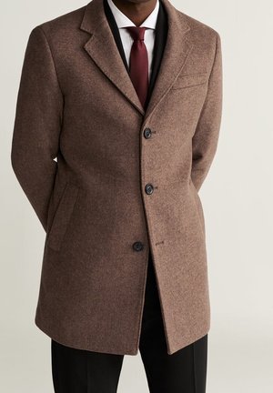 Bruine wollen overjas met schouderklappen, drie zwarte knopen en zijzakken, gedragen over een wit overhemd en een bordeauxrode stropdas.
