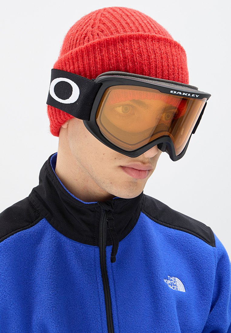 Czarne gogle narciarskie z pomarańczowymi soczewkami noszone przez osobę w niebieskiej polarowej kurtce, dopełnione czerwoną wełnianą czapką. Widoczna wyraźna marka logo.