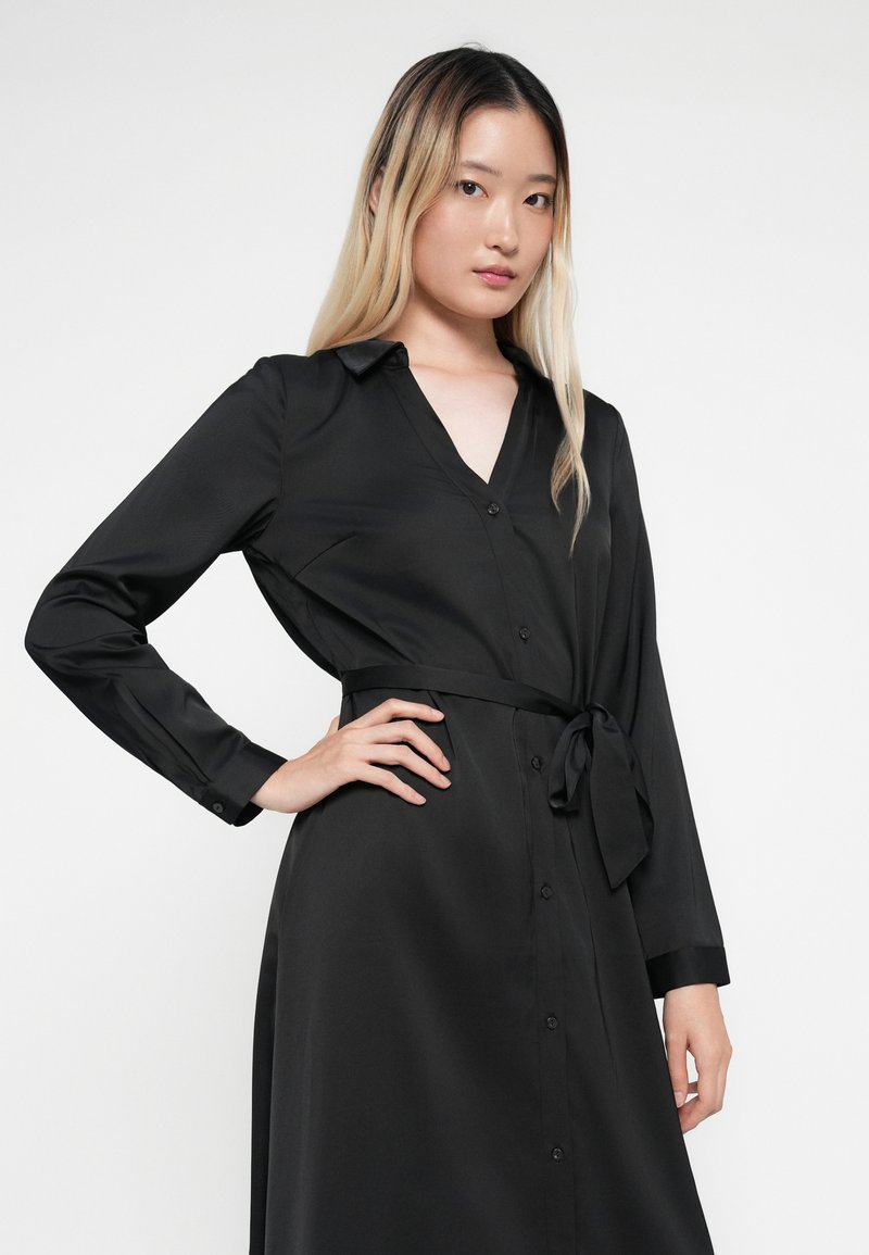 Zwarte blouse jurk van een gladde stof, met een kraag, lange mouwen en een gestrikte riem in de taille.