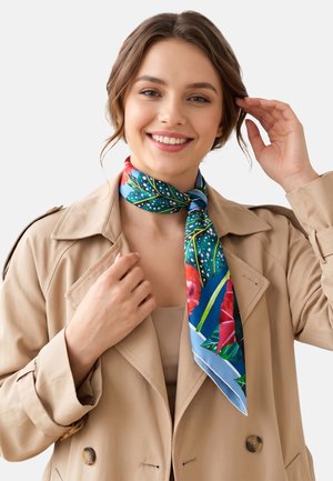 Joven sonriente, vistiendo un abrigo beige y una bufanda floral colorida atada alrededor del cuello, ajustándose el cabello con una mano.