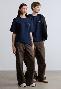 Μπλουζάκια από βαμβάκι σε ναυτικό μπλε με μικρό πράσινο λογότυπο, συνδυασμένα με καφέ παντελόνια σε άνετη γραμμή και αθλητικά παπούτσια, τοποθετημένα σε ουδέτερο φόντο.