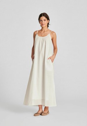 Naine, kes kannab pikka, varrukateta valget kleiti õhukeste rihmade ja taskutega, seisab taskutes käed, jalas beežid madalad kingad heledal hallil taustal.